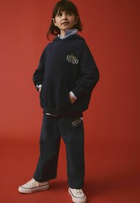 Ensemble de sweat à capuche et de pantalon de jogging bleu marine avec des détails de logo, offrant une coupe décontractée et une texture douce. Des baskets blanches complètent la tenue.