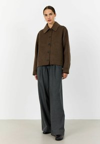 Woman standing wearing a brown buttoned jacket, wide-leg gray trousers, black shoes, and gold drop earrings against a plain white background.