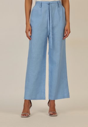 Frau trägt weite, hellblaue, hoch taillierte Hose mit Stoffgürtel und silbernen offenen Sandalen, steht vor einem beigen Hintergrund.