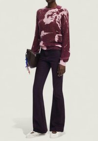 Fioletowy sweter tie-dye z długimi rękawami, połączony z ciemnofioletowymi spodniami dzwonami i białymi płaskimi butami. Brązowa torebka z kolorowymi akcentami.