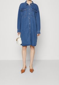 Denim shirt dress in light blue with long sleeves, two chest pockets, button-down front, and a relaxed fit. Paired with pointed brown flats.