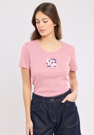 Femme portant un t-shirt rose avec un petit motif de feuilles bleu et blanc sur la poitrine et un pantalon en denim sombre taille haute.