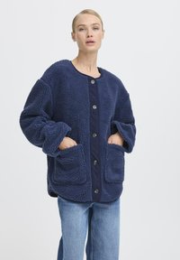 Veste en polaire bleu marine avec une surface texturée, un col rond, de grandes poches plaquées et des boutons noirs sur le devant.
