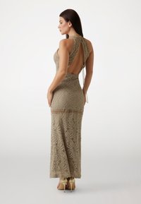 Lace maxi dress in beige with an open back, featuring a fitted shape, intricate floral pattern, and side slit details. Paired with high heels.