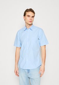 Camisa de botones de manga corta color azul claro con un bolsillo en el pecho, diseño casual, cuello con solapa y detalles de costura visibles.