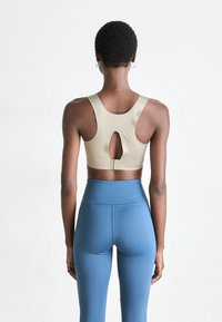 Kvinne med kort, krøllete hår iført en beige racerback sportsbh og høyhåndte blå tights, vendt mot en enkel hvit bakgrunn.