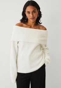Off-the-shoulder hvid sweater med ribbet tekstur, lange ærmer og en afslappet pasform. Sammen med sorte bukser, der viser et afslappet look.