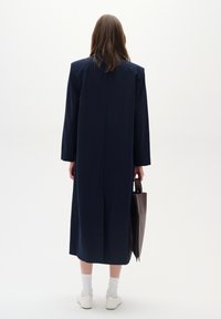 Cappotto lungo blu navy con taglio dritto e spalline, dotato di maniche lunghe e cucitura centrale sul retro. Il modello porta una borsa scura.