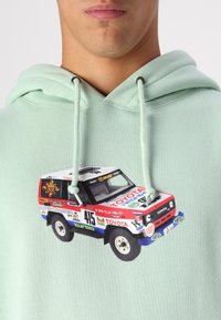 Mátově zelená mikina s grafikou Toyoty Land Cruiser v červenobílém designu, zobrazující různé loga a čísla.