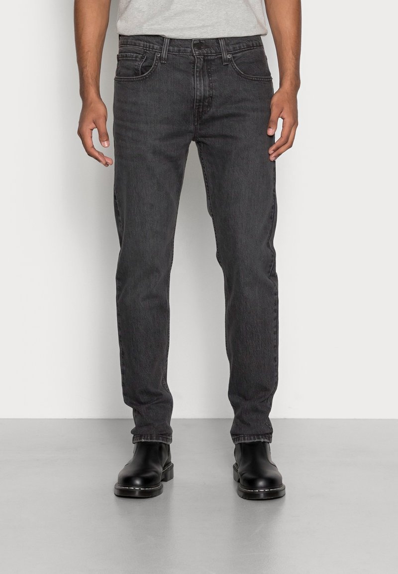 Homme portant un jean noir à jambes droites et des bottes en cuir noires, debout sur un sol gris contre un mur blanc.