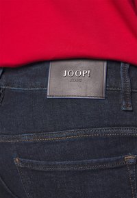 Mörkblå denimjeans med en läderbakpanel som har den präglade logotypen "JOOP! JEANS." Synlig sömnad i en kontrasterande färg.