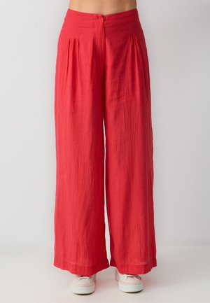 Rote, weit geschnittene Hosen aus leichtem Stoff mit Falten an der Taille und einem Knopfverschluss an der Rückseite. Getragen mit weißen Turnschuhen.