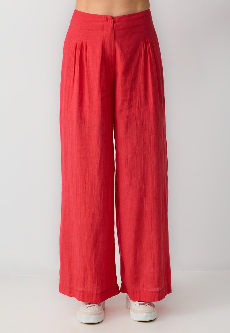 Pantalons rouges à jambes larges en tissu léger, avec des plis à la taille et une fermeture à bouton à l'arrière. Portés avec des baskets blanches.