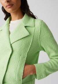 Giacca in misto lana verde chiaro con colletto a revers, due tasche frontali e chiusura a bottone. Tessuto testurizzato con cuciture visibili.