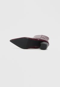 Stivale alla caviglia bordeaux a punta con texture coccodrillo e tacco a blocco, mostrato dal lato della suola su uno sfondo bianco.