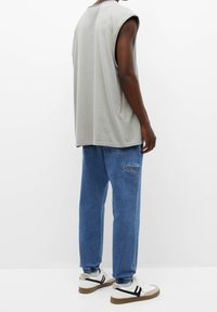 Persoon in een mouwloos grijs shirt, blauwe joggingjeans met zakken en witte sneakers met zwarte strepen, staand voor een witte achtergronden.