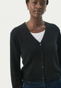 Čierny cardigan vyrobený z jemného úpletu, s V-výstrihom, dlhými rukávmi a štyrmi gombíkmi vpredu, nosený cez biely top.