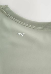 Šviesiai žalia sportinė palaidinė su apvaliu iškirpimu. Pasižymi lygiu, tampriu tekstūru ir mažu baltu „self.“ logotipu netoli apykaklės.