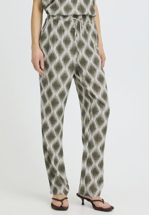 Femme portant un pantalon ample à motif ikat vert et blanc avec une taille nouée, associé à des sandales à talons marron, debout devant un fond uni.