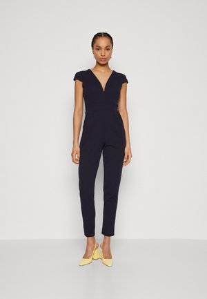 Marineblå jumpsuit med V-udskæring og korte ærmer, med tætsiddende ben og defineret talje. Sammen med gule hæle.
