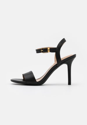 Black high-heeled sandal with open toe, ankle strap, and gold buckle detail, set against a white background.