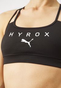 Crna sportska brusnica od glatkog materijala s širokim naramenicama, niskim izrezom i bijelim tekstom "HYROX" s Puma logom u sredini.