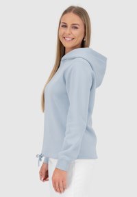 Felpa leggera blu chiaro con coulisse all'orlo e maniche a polsino, che presenta una superficie liscia e una vestibilità casual, indossata con pantaloni bianchi.