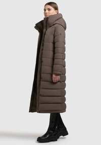 Piumino lungo marrone scuro, con collo alto e design trapuntato orizzontale. Abbinato a stivaletti neri alla caviglia e leggings.