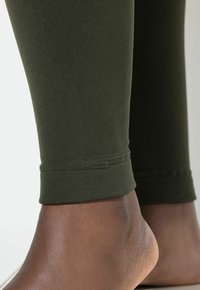 Olivegrüne Leggings aus einem glatten, dehnbaren Stoff, mit einem enganliegenden Knöchelbund und einem nahtlosen Design entlang des Unterschenkels.