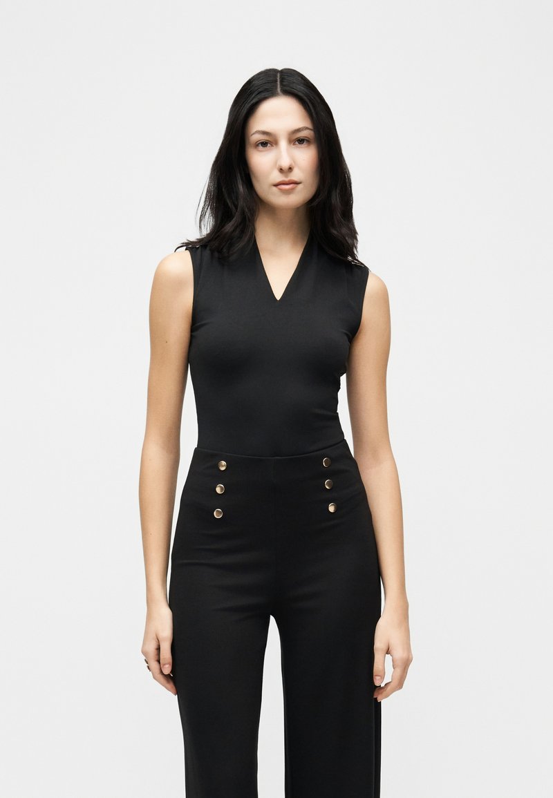 Γυναίκα με μακριά σκοτεινά μαλλιά, φορώντας ασύμμετρο μαύρο τοπ χωρίς μανίκια και μαύρο μπλουζάκι με ψηλά κουμπιά χρυσού χρώματος, στέκεται μπροστά από απλό φωτεινό φόντο.