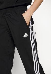 Czarne sportowe spodnie wykonane z lekkiego materiału, z białymi paskami po bokach i wyszywanym logo. Elastyczna talia z bocznymi kieszeniami.