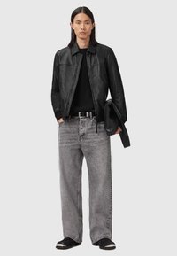 Man som bär en svart läderjacka över en svart tröja, grå vida jeans, svart bälte och svarta skor, håller en hopvikt svart jacka.