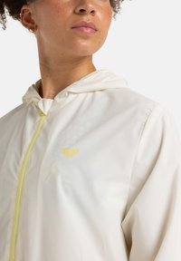 Lightweight hooded jacket in beige, featuring a yellow zipper and logo. Smooth texture with subtle patterned detailing.