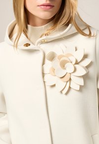 Weißer Wollmantel mit Kapuze, ausgestattet mit einer großen geschichteten Blumenapplikation in Creme und Beige sowie goldfarbenen Knöpfen auf der Vorderseite.