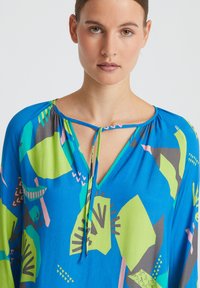 Haut à motif bleu et vert avec un décolleté en V et un détail de nœud. Tissu fluide présentant des motifs géométriques et de feuilles en rose et gris.