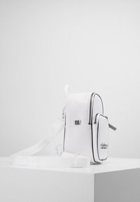 Malá bílá crossbody taška Adidas s černým logem, přední kapsa na zip, nastavitelné ramenní popruhy, vystavena na bílé podložce na jednobarevném pozadí.