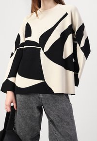 Pull beige et noir à motifs, coupe ample, encolure ronde et manches larges, associé à un pantalon gris et un sac noir.