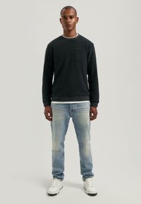 Uomo in piedi di fronte, indossa una maglietta nera a maniche lunghe, jeans azzurri chiari sbiaditi e sneaker bianche, su uno sfondo neutro.