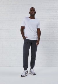 Mann trägt ein weißes kurzärmeliges T-Shirt, dunkelgraue Jogginghose und weiße Sportschuhe und steht vor einer weißen Ziegelwand.