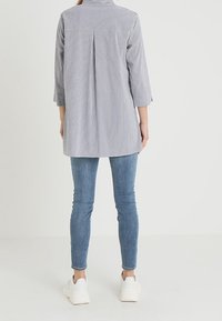 Koszula w paski z kołnierzykiem, rękawami do 3/4 długości i zakładką z tyłu, zestawiona z niebieskimi obcisłymi dżinsami i białymi trampkami. Jednolite tło.