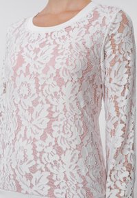 Λευκό τοπ από δαντέλα με ανθισ�μένο σχέδιο, στρογγυλή λαιμόκοψη και μακρύ μανίκι. Διαθέτει ημι-διάφανη υφή πάνω σε ελαφρώς ροζ βάση.