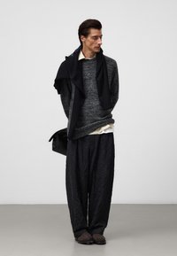 Sweter w ciemnoszarym splocie, nałożony na białą koszulę z kołnierzykiem; szerokie, fakturowane czarne spodnie oraz czarne buty wsuwane, trzymający czarną torbę.