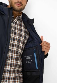Uomo che indossa una giacca blu navy aperta per mostrare la tasca interna con zipper azzurro e etichetta di isolamento Thermore, sopra una camicia a quadri marrone e blu.