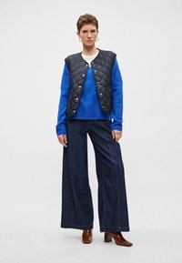 Fekete steppelt mellény ezüst patentokkal, élénk kék pulóver fölött, széles szárú sötét farmernadrággal és barna bokacsizmával párosítva.