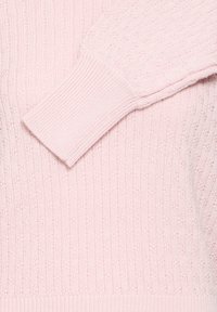 Pull en tricot rose clair avec poignets côtelés, présentant un motif texturé et des détails en relief sur l'ensemble du tissu. Design simple et confortable.