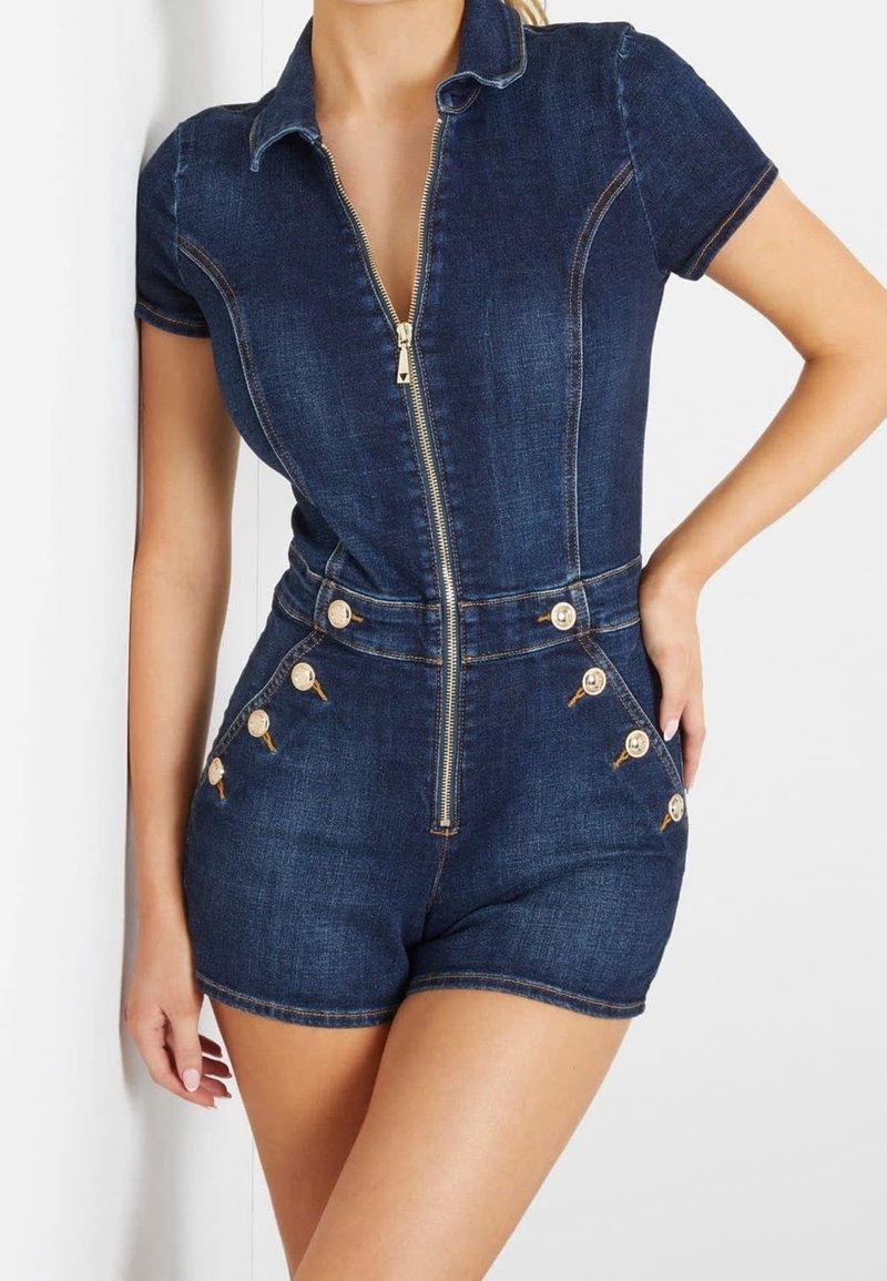 Femme portant une combinaison courte en denim bleu foncé à manches courtes avec une fermeture éclair sur le devant et des boutons dorés sur les côtés, debout avec une main sur la hanche.