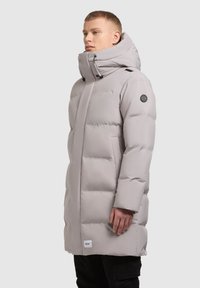 Garšs, spilventots mētelis gaiši pelēkā krāsā ar augstu apkakli, sānu kabatām un melnu logo uzlīmi uz piedurknes. Gluda, izolēta tekstūra.