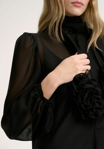 Blusa negra con tejido transparente, cuello alto y mangas abullonadas voluminosas. Presenta un acento con volantes en la parte delantera. Textura suave en general.