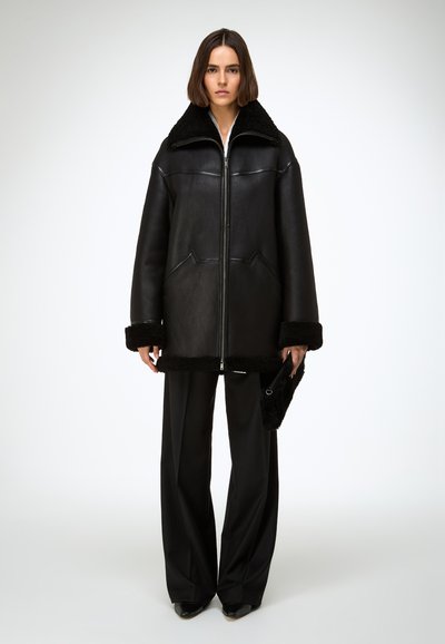 Manteau en cuir noir avec col et poignets en peluche, fermeture éclair et détails de couture subtils. Associé à un pantalon noir à jambes larges.