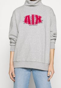 Felpa grigia con collo alto, caratterizzata da un audace logo "AIX" in rosa e testo nero. Vestibilità oversize con polsini e orlo a costine. Abbinata a jeans azzurri.